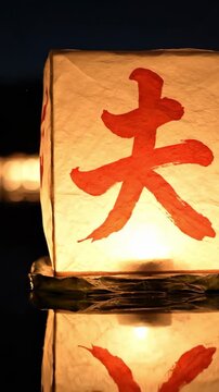 Single washi paper Obon lantern glowing gold on still black water at night, perfect reflection below, distant bokeh lanterns background, Obon tōrō nagashi ancestor floating lantern Japan sacred scene.