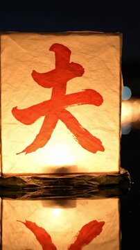 Single washi paper Obon lantern glowing gold on still black water at night, perfect reflection below, distant bokeh lanterns background, Obon tōrō nagashi ancestor floating lantern Japan sacred scene.