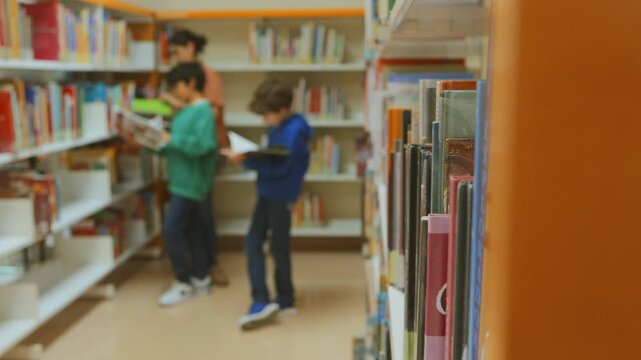 Children reading books in a public library, with a woman guiding them through the bookshelves. Boys and mother choosing literature and reading stories in a bookstore aisle