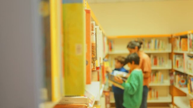 Unfocused mother and two young sons choosing a book together in a library, selecting literature from a bookshelf, representing family learning and childhood education