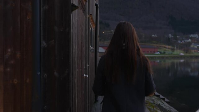 Woman walks beside wooden fjordside saunas by calm reflective water in Skjolden Norway
