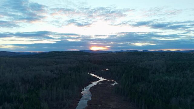 Winding northern New Hampshire river at sunset, a high-altitude aerial drone landscape showcasing a serpentine river cutting through a dense, vast evergreen forest under a dramatic cloudy sky at dusk.