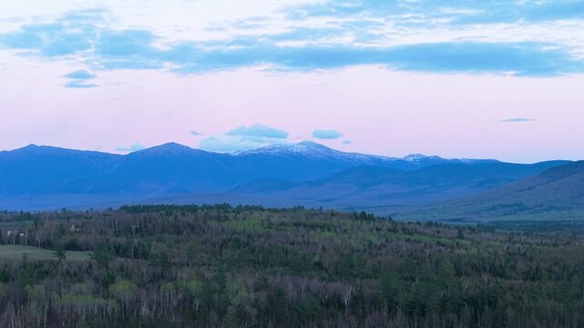 White Mountains northern New Hampshire sunset landscape, aerial drone video over vast pine forests looking toward distant blue mountain peaks with snow-dusted summits under a twilight sky.