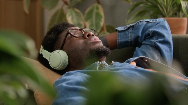 Handheld slowmo shot of young Black man wearing headphones resting on couch and listening to music, surrounded by green houseplants providing tranquility and stress relief