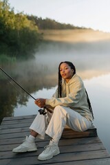 Fototapeta premium Young woman fishing on dock by misty lake at sunrise