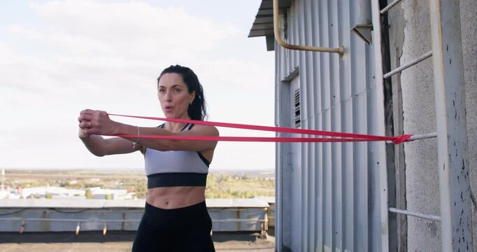 Sportive effort. Outdoor resistance training. Motivated female athlete performing intensive practice extending red band on rooftop.