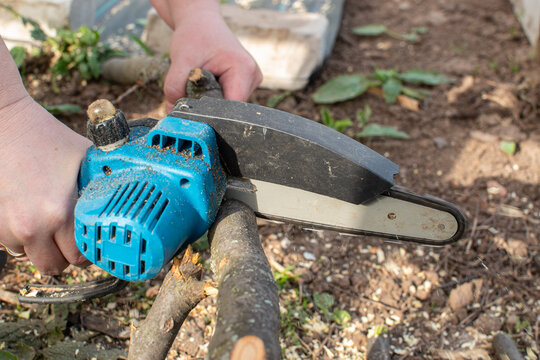 Close-up of a person using a cordless mini chainsaw to cut dry tree branches on the ground in the backyard. Gardening and yard work concept