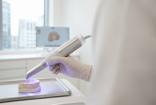 Gloved hand holds handheld dental 3D intraoral scanner device scanning plaster tooth model inside modern dentistry clinic room. Healthcare hospital support. Medical diagnostic equipment. Copy-space.