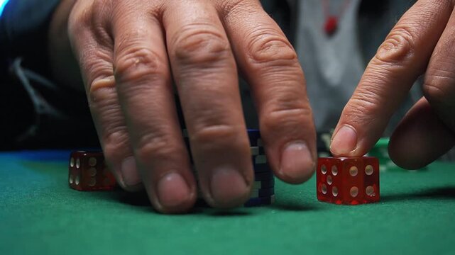 Close-up of hands placing red dice on green felt table with stacked poker chips and additional dice in a dimly lit gaming environment