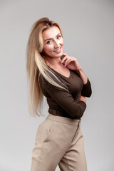 Fototapeta premium Graceful middle aged woman touching neck while posing sideways with gentle smile wearing brown long sleeve shirt and beige trousers standing isolated over gray background