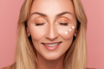 Naklejka premium Mid-adult blonde woman smiling gently with closed eyes and three moisturizer dots under eye during skincare isolated on pink background