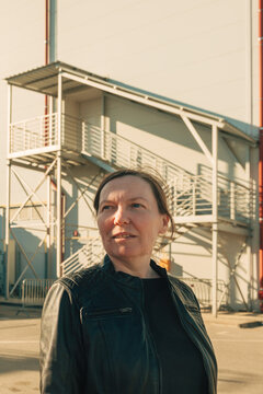 Confident middle-aged woman in a leather jacket stands outside an industrial building at golden hour, looking thoughtful.
