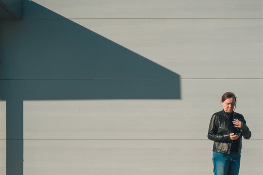 Woman in a black leather jacket stands against a minimal pale wall, looking at her mobile phone as a strong geometric shadow falls across the facade, evoking solitude and modern life.