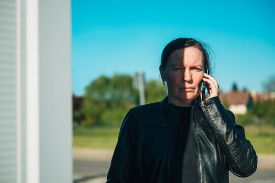 Serious middle-aged woman standing outdoors in a suburban area, wearing a black leather jacket while making a phone call on a mobile device under bright sunlight.