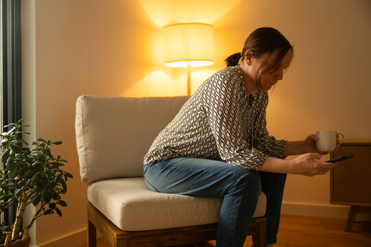 Mature woman checking smartphone with cup of coffee indoors, cozy communication and everyday routine concept