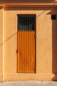 Orange metal door set into a textured stucco wall, bathed in warm sunlight and long shadows. Minimal urban architecture with bold color, geometric lines and a quiet, summery mood.
