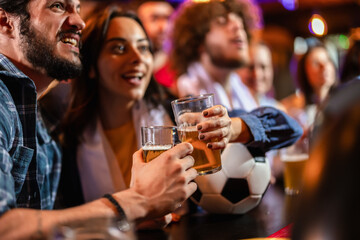 Two sports fans toasting with beer next to a soccer ball at a pub bar counter.