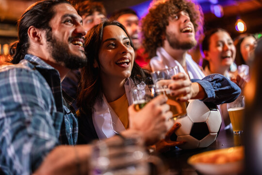 Two friends toasting with beer by a soccer ball with people cheering in the background.