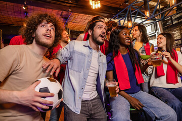 Fototapeta premium Happy group of soccer fans with red scarves smiling and watching a match in a pub.