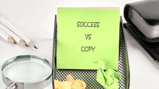 Person writes the phrase Success versus Copy on a bright green sticky note within a black wire desk organizer. Pencils, a magnifying glass, and crumpled paper are nearby