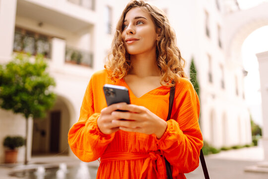 A woman stands on a city street wearing an orange dress. She holds a smartphone and appears to be focused on the screen. Concept of style, fashion, and walking.