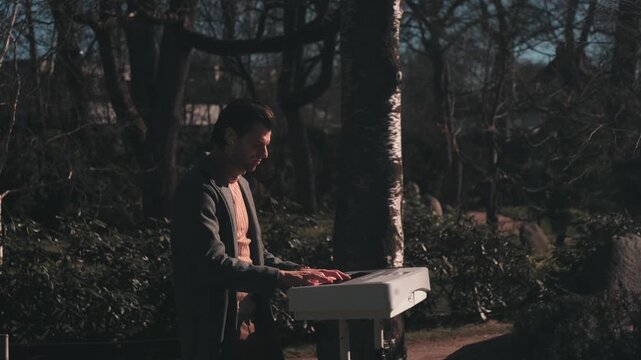 Atmospheric cinematic scene of young male musician playing white digital piano outdoors in forest park lighting.