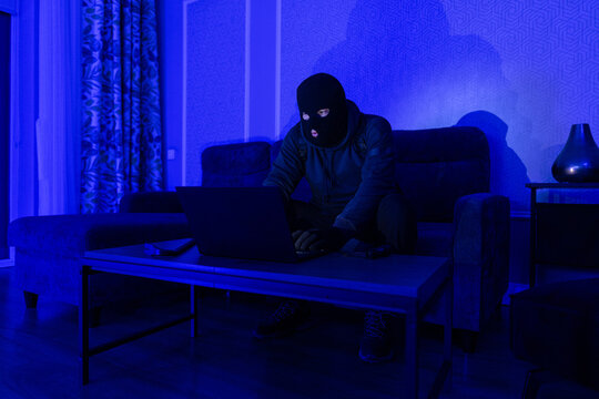 Cybercrime and spying concept. Masked man criminal using laptop computer at night, making cyberattack, breaking the law, sitting in dark room