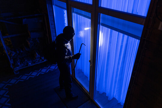 Man with crowbar standing outside looking through window to break into house at night, above view shot