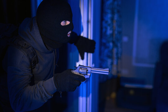 Man thief in mask holding gun while entering dark room at night through balcony door, looks ready to commit robbery