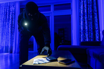 Fototapeta premium Home invasion. Masked robber breaking into house at night through door, using flashlight and taking valuables, cash and smartphone from table