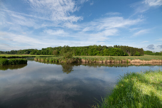 A calm river flows through open fields. The river splits into two branches. The sky is reflected in the water.