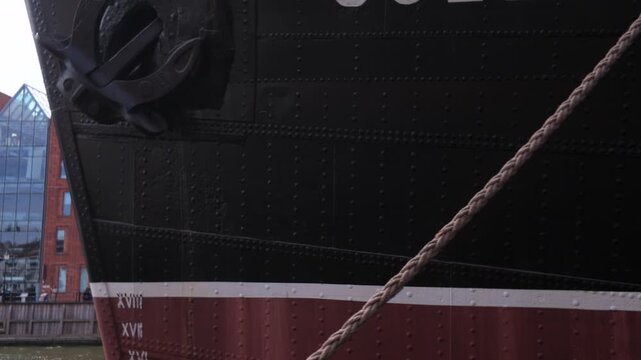 Close-up of a vintage cargo ship bow with a steel anchor and draft marks, set against historic brick waterfront buildings, illustrating maritime logistics.