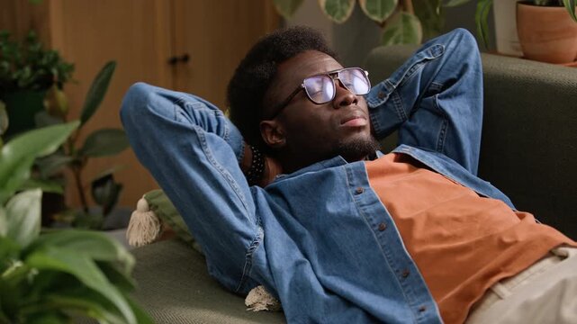 Medium slowmo shot of young African American man wearing denim shirt and eyeglasses relaxing on couch among green houseplants and enjoying tranquility while practicing mindfulness at home