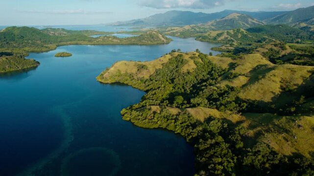 Drone shot pulling back and tilting down over green savanna hills and tropical blue sea bays in Riung 17 Islands National Park, Flores.
