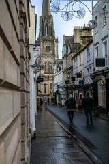 Naklejka premium View of the Gothic Revival tower of St Michael's Church overlooking a wet cobblestone street in Bath. The historic church tower rises above the Georgian cityscape, with rain-slicked streets