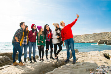 Obraz premium Group of friends taking selfie on rocky beach.Seven young adults, smiling and laughing, pose for a photo on a rocky coastline