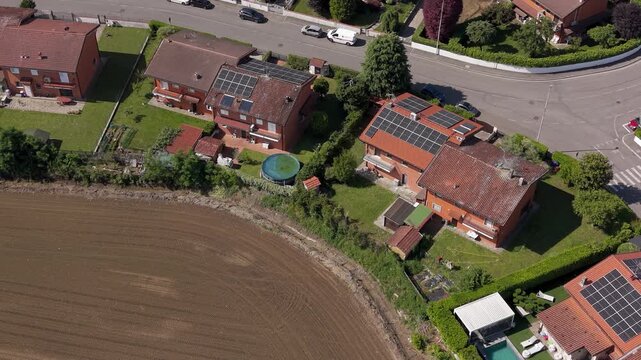 Aerial View of Suburban Homes: A high-angle shot reveals a cluster of residential houses with tiled roofs and solar panels, nestled amidst green gardens and bordering a field.