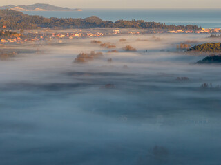 Naklejka premium Serene morning aerial scenery of a misty valley in North Corfu Greece at golden hour sunrise showing thick white fog covering green fields, trees, local houses and the coastline.