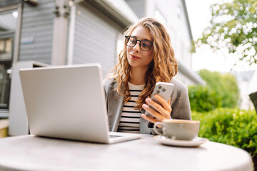 Fototapeta premium Woman sits at table outside cafe, smiling and using phone. Laptop and a cup of coffee are on table. Female freelancer works on terrace, phone in hand. Concept of remote work, technology.