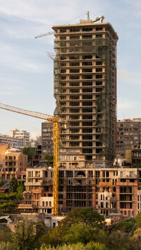 Modern concrete residential or commercial tower skyscraper under construction covered in safety nets with a yellow tower crane in the foreground