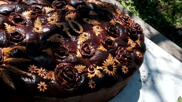 A large traditional korovai bread baked for a 70th anniversary is displayed outdoors in bright sunlight. The number 70 is formed in the center, surrounded by decorative dough .