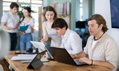 Fototapeta premium Adult man at desk in modern office and working on laptop among group of colleagues