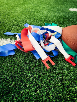 Vertical close-up of a brown football resting next to a pile of red, blue, and white flag football belts on green athletic turf. Great for mobile-first sports content.