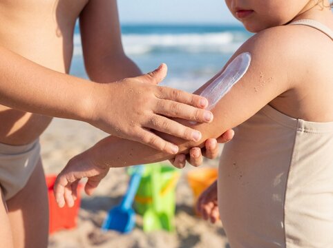 Primer plano de un ni&ntilde;o aplicando protector solar a su hermano menor en una playa con juguetes difuminados al fondo.