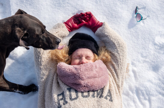 Woman lying on snow with eyes closed wearing winter clothing while a dog licks her face creating a tender playful moment of trust warmth companionship and joy in cold nature
