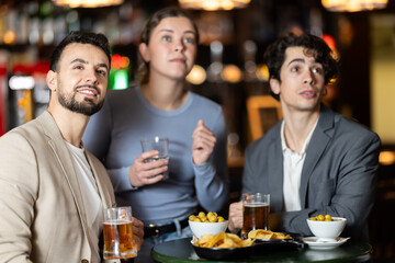 Fototapeta premium Group of excited young friends fans cheering for sports team together, watching match on TV in big beer bar
