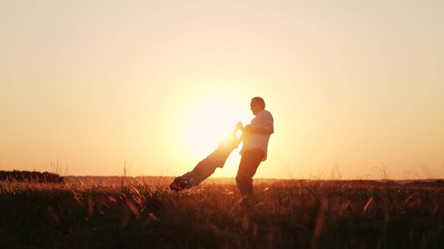 Dad plays with child, holding son hands, twirls him around, sunset field. Family picnics outdoors. Dad plays with son in summer park. Childhood dream fly. Father, child boy, twirling in sunny park.