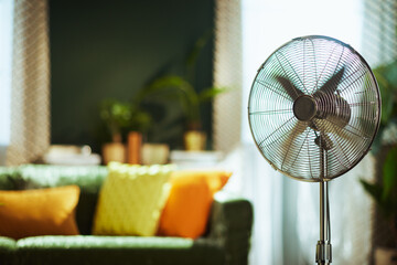 A modern stand fan with spinning blades is prominently featured, providing refreshing air circulation in a comfortable living room with a green velvet sofa and natural light, ideal for heat relief. © Alliance