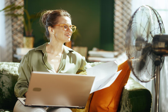 An adult woman with orange glasses smiles, eyes closed, enjoying the cool breeze from a fan. On a green sofa with laptop & papers, she embodies comfort & productivity in a warm home office setting