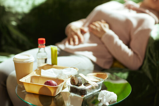 Lying on a sofa, a woman rests her hands on her bloated stomach after eating takeout. Sunlight filters through as she recovers from the discomfort of a heavy, unhealthy meal.